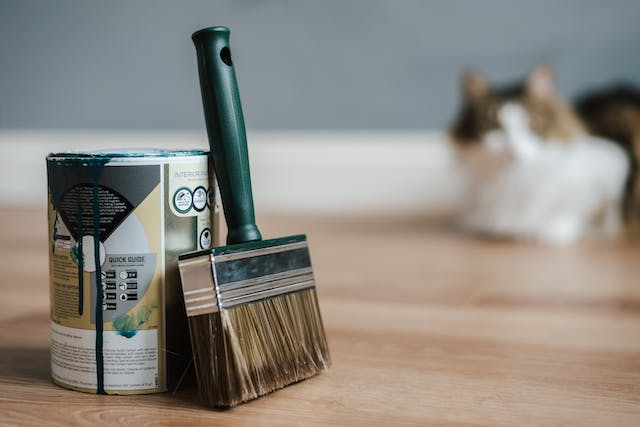 A can of paint and a large brush on the floor with a cat in the background.